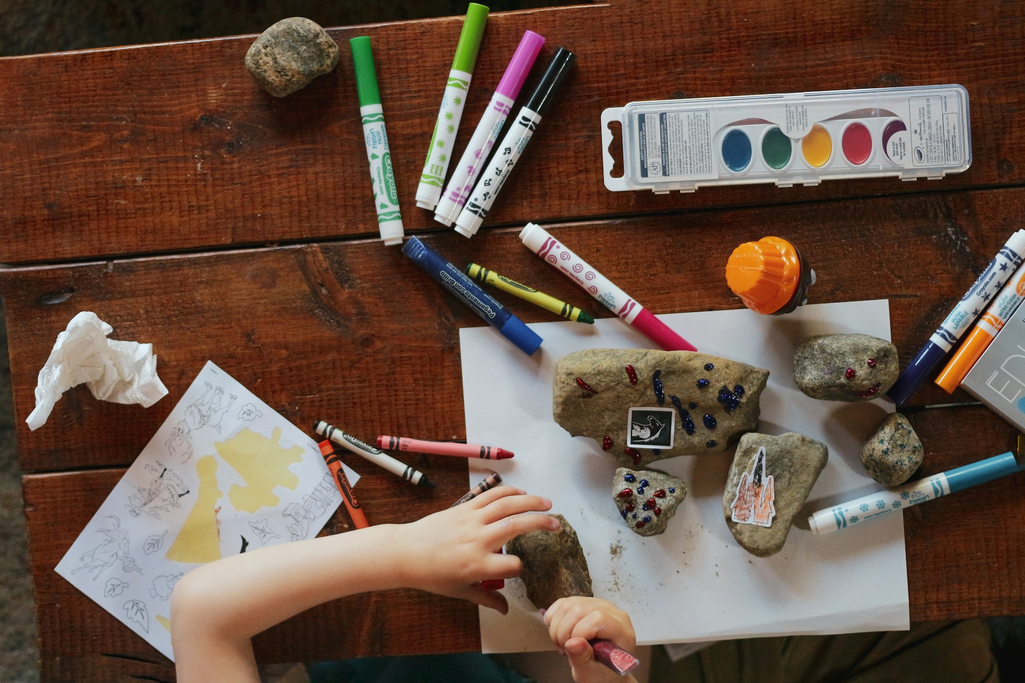 A child's hands painting and decorating rocks with markers and watercolours on a wooden table